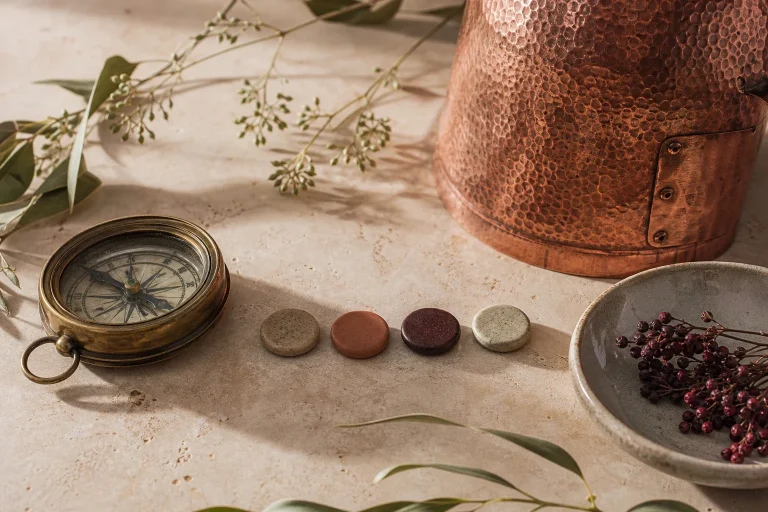 Nature morte d’atelier avec un alambic en cuivre, une boussole, quatre pastilles colorées et des végétaux sur une table.